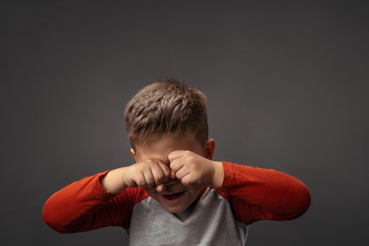 Crying little boy expresses sad emotions. Upset child rubs eyes with fists isolated on gray background. Conflict concept.