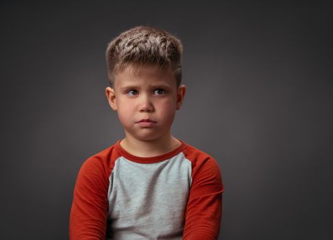 Sad boy looking at camera. Preschool kid expresses emotions. Isolated on gray background. Conflict concept. Copy space at right.