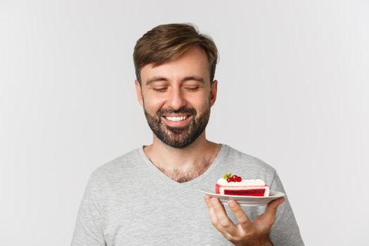 Close-up of handsome bearded man, smiling and looking at tasty cake, standing over white background.