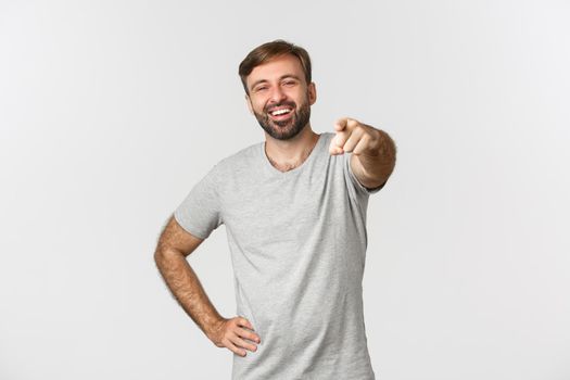 Image of carefree caucasian guy with beard, wearing gray t-shirt, laughing and pointing finger at camera, standing over white background.