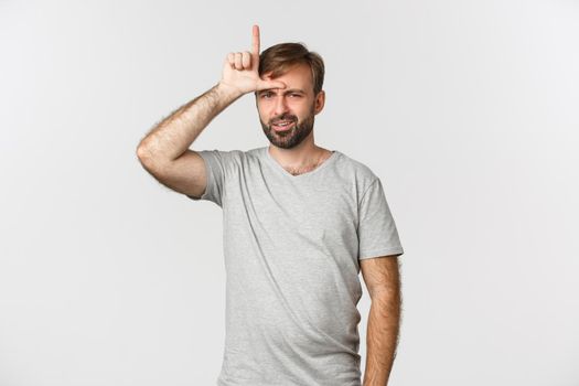 Portrait of arrogant man in gray t-shirt, mocking person who lost, showing loser sign on forehead and looking with dismay, standing over white background.