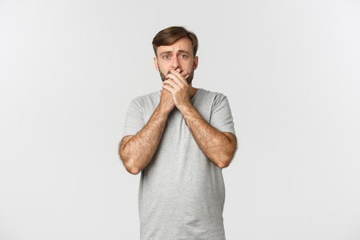 Image of scared man in gray t-shirt, gasping and shivering from fear, standing anxious over white background.
