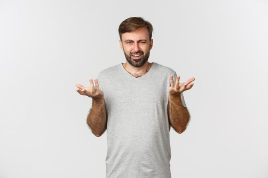 Portrait of disappointed and confused man with beard, raising hands up and complaining, standing in gray t-shirt over white background.