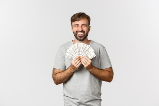 Happy man smiling pleased, showing money, standing over white background in gray t-shirt.