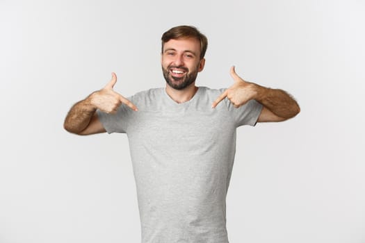 Portrait of handsome bearded man in gray t-shirt pointing at logo and smiling, recommending brand, standing over white background.