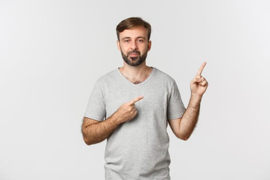 Image of handsome bearded guy in gray t-shirt, showing logo, pointing fingers at upper right corner, standing over white background.