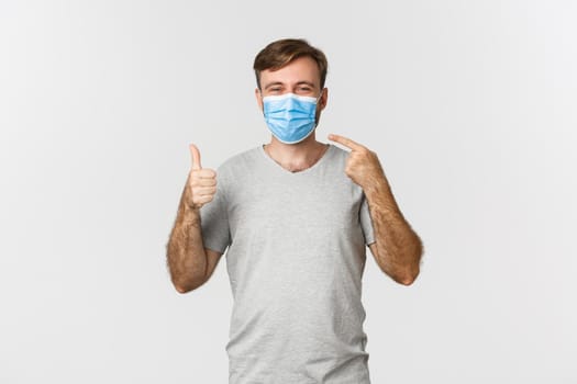 Concept of pandemic, covid-19 and social-distancing. Image of happy guy in gray t-shirt, pointing at medical mask and showing thumbs-up, standing over white background.