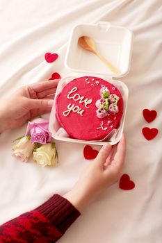 Womens hands are holding a plastic lunch box with a wooden spoon and a delicious red bento cake with the inscription Love you, with red hearts and roses lying on a white bed next to it. View from above. Vertical.