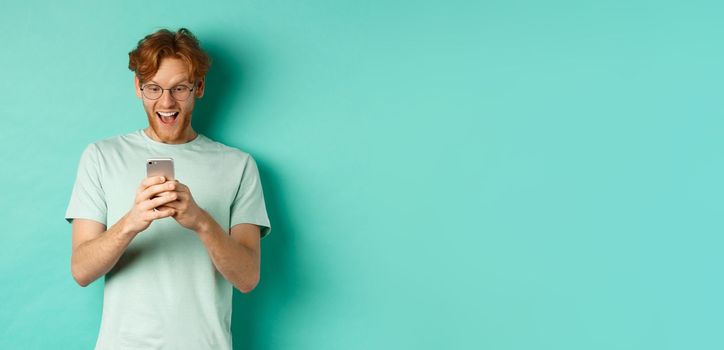 Image of young redhead man in glasses reading phone screen with surprised face, receive amazing promo offer, standing over turquoise background.