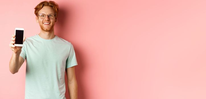 Handsome redhead man in glasses showing blank smartphone screen and smiling, demonstrate online promo or application, standing over pink background.