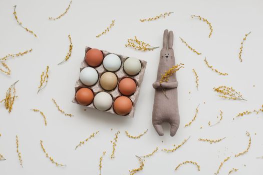 Funny handmade bunny rabbit toy with colorful chicken eggs and yellow mimosa flowers. Easter gift concept.