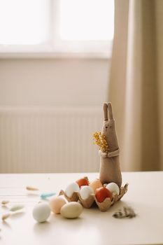 Funny handmade bunny rabbit toy with colorful chicken eggs and yellow mimosa flowers. Easter gift concept.