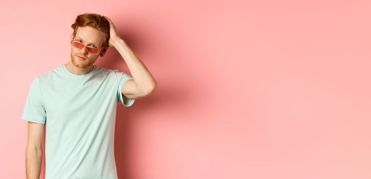 Handsome young redhead man in sunglasses, brushing hair with hand and looking smug and confident at camera, standing over pink background.