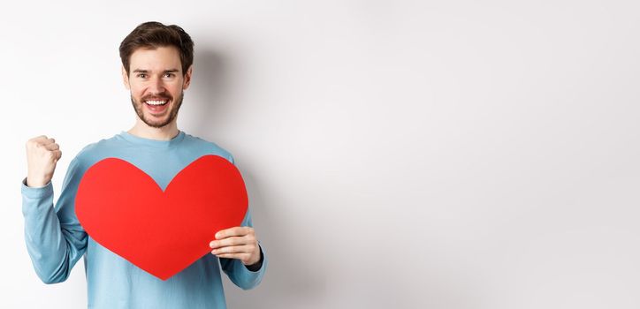 Cheerful man determined to find love on Valentines day, making fist pump success gesture and holding big red romantic heart, standing over white background.