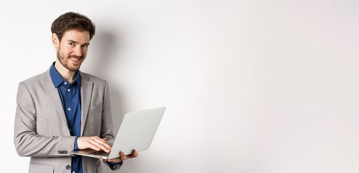 Successful smiling businessman working on laptop and looking happy at camera, standing in grey suit on white background.