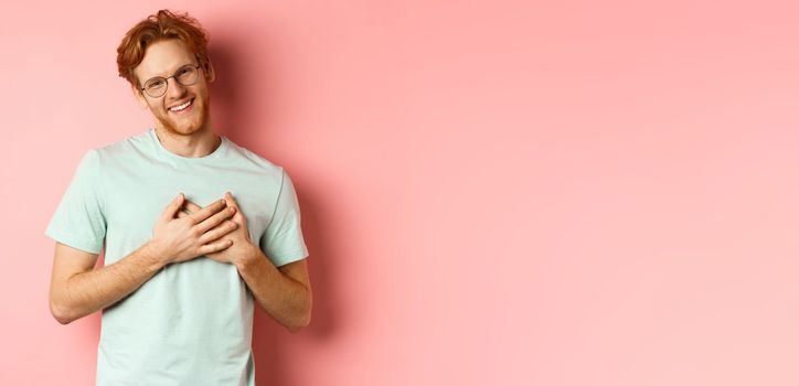 Attractive young man with ginger hair, holding hands on heart and smiling grateful, saying thank you, express gratitude, standing over pink background.