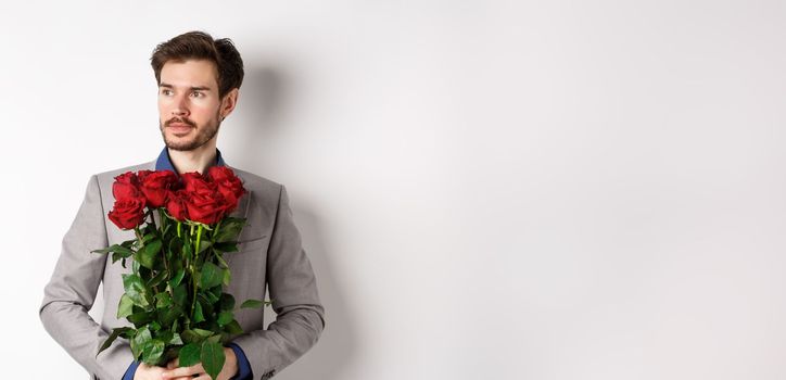 Handsome boyfriend in suit going on romantic date, holding bouquet of red roses and looking left thoughtful, standing over white background.