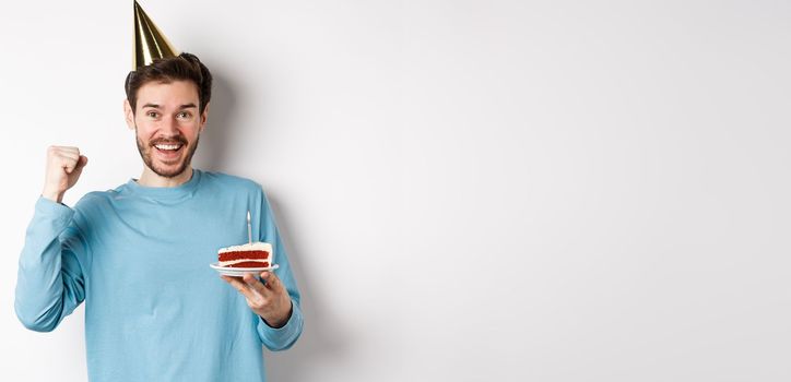 Celebration and holidays concept. Cheerful young man celebrating birthday in party hat, holding bday cake and looking happy, standing on white background.