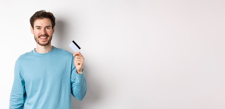 Smiling male bank client showing plastic credit card with happy face, standing over white background.