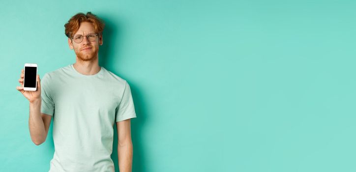 Skeptical male model with red hair and glasses, showing mobile screen and frowning disappointed, dislike application, standing over turquoise background.