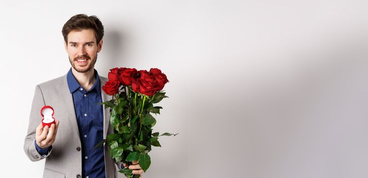 Smiling boyfriend making a marriage proposal, standing with engagement ring and bouquet of red roses, going on romantic Valentines day date, white background.