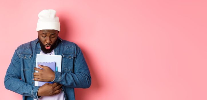 Education. Image of african-american bearded male student holding notebooks and looking down, drop something on floor, standing over pink background.