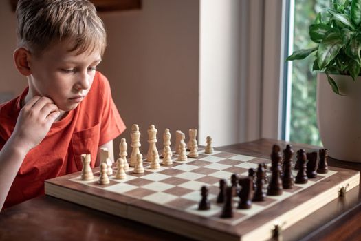 Young white child playing a game of chess on large chess board. Chess board on table in front of school boy thinking of next move, tournament