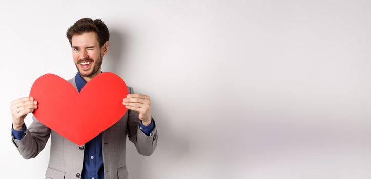 Charismatic young man winking and smiling, showing big red heart cutout for Valentines day date, say love you to lover, standing over white background.