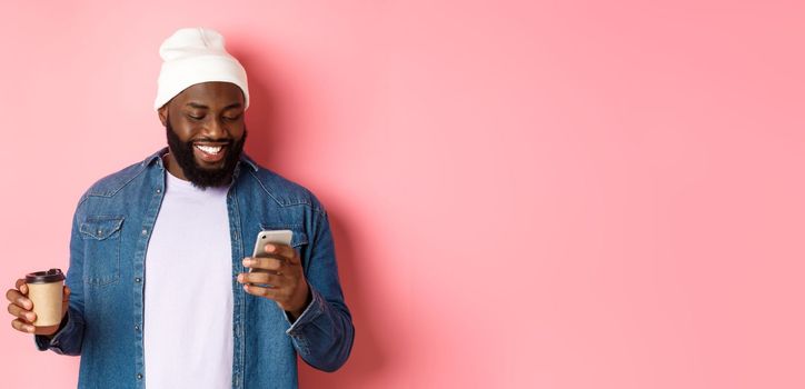 Image of stylish Black man hipster drinking takeaway coffee, reading message on phone and smiling, standing over pink background.