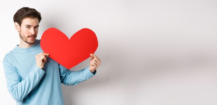 Valentines day. Handsome and romantic man holding big red valentine heart cutout, looking seductive at camera, making love confession, white background.