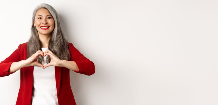 Beautiful asian mature woman in red blazer and makeup, showing heart sign and smiling, I love you gesture, standing over white background.