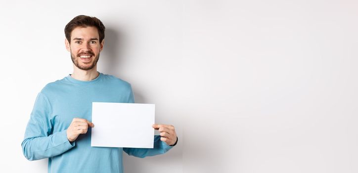 Young man with happy face showing blank piece of paper for your logo or sign, smiling at camera, standing over white background.