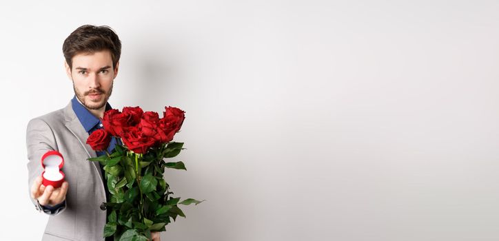 Handsome young man making a marriage proposal, stretch out hand with engagement ring and holding red roses, asking to marry him, looking confident at lover, white background.