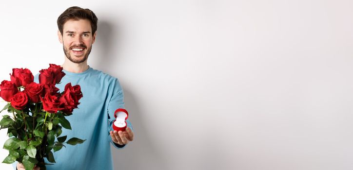 Handsome young man boyfriend making a proposal on Valentines lovers day, holding bouquet of red roses and engagement ring, concept of wedding and relationship.