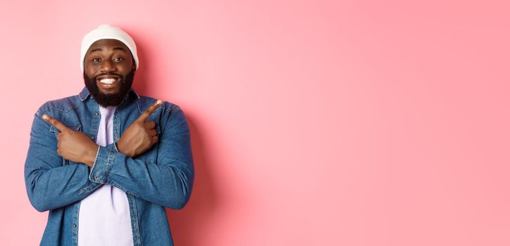 Happy young african-american hipster guy pointing fingers sideways, smiling and showing two choices, showing offers, standing over pink background.