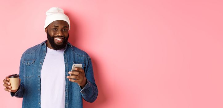 Image of stylish Black man hipster drinking takeaway coffee, reading message on phone and smiling, standing over pink background.