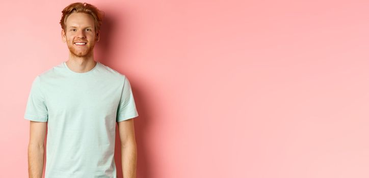 Happy redhead man in t-shirt looking at camera and smiling, standing over pink background.