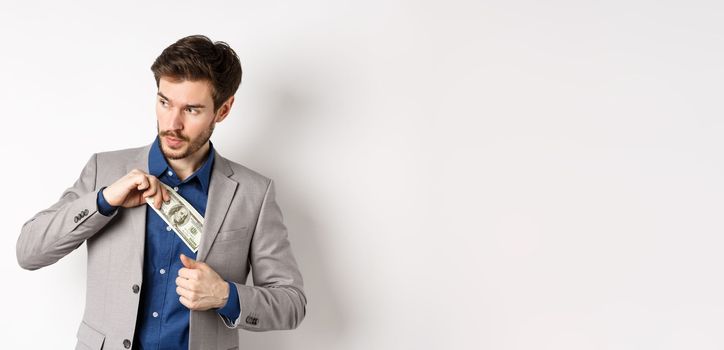 Confident businessman put money in suit and looking aside, standing on white background.