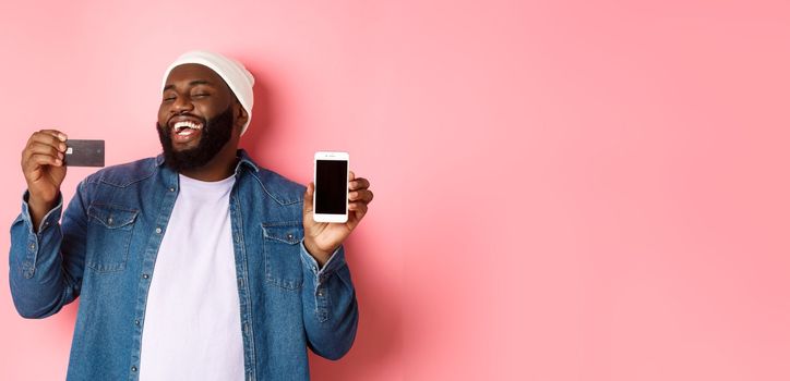 Online shopping. Happy african-american man in beanie laughing, showing credit card and mobile phone screen, standing over pink background.