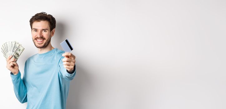 Smiling caucasian man holding money and giving you plastic credit card, standing on white background.