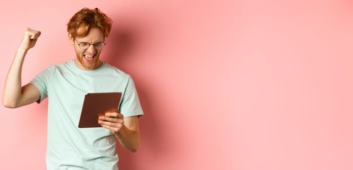 Image of happy redhead man triumphing, winning online with digital tablet and rejoicing, standing over pink background.
