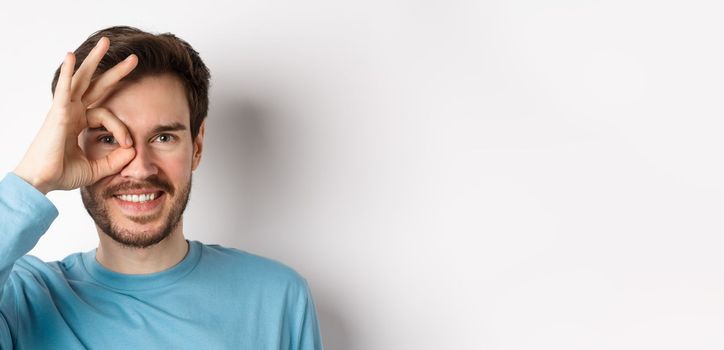 Close up of happy man looking through okay sign and smiling, approve and like something good, standing over white background.