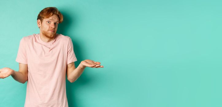 Young redhead guy in t-shirt know nothing, shrugging shoudlers with hands spread sideways, standing confused over turquoise background.