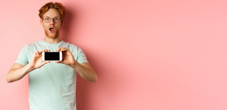 Amazed redhead man gasping and staring with awe at camera, showing blank smartphone screen horizontally, standing over pink background.