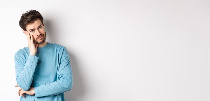 Sad young man looking frustrated, touching temple of head and frowning uneasy, standing against white background.