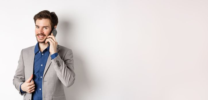 Successful businessman fixing his suit and talking on mobile phone, smiling at camera, standing on white background.