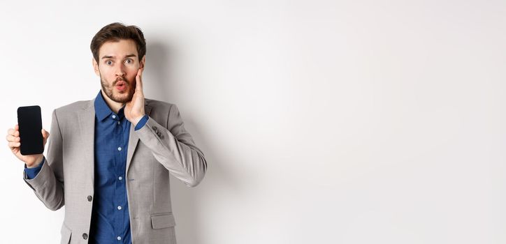 E-commerce and online shopping concept. Excited man gasping amaze and showing empty smartphone screen, wearing business suit, white background.