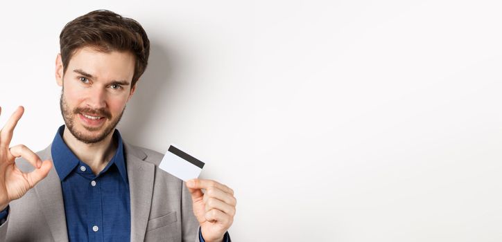 Shopping. Handsome man showing okay sign and plastic credit card, all under control, no worries gesture, white background.