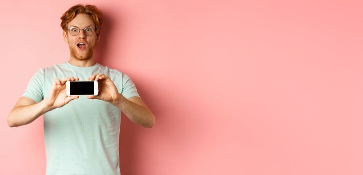 Amazed redhead man gasping and staring with awe at camera, showing blank smartphone screen horizontally, standing over pink background.