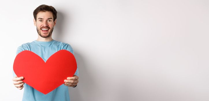 Relationship and people concept. Handsome man give valentines heart to you, smiling and saying I love you at camera, standing over white background.